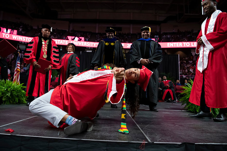 Administrators stand behind drum major in cap and gown as he performs on stage at commencement