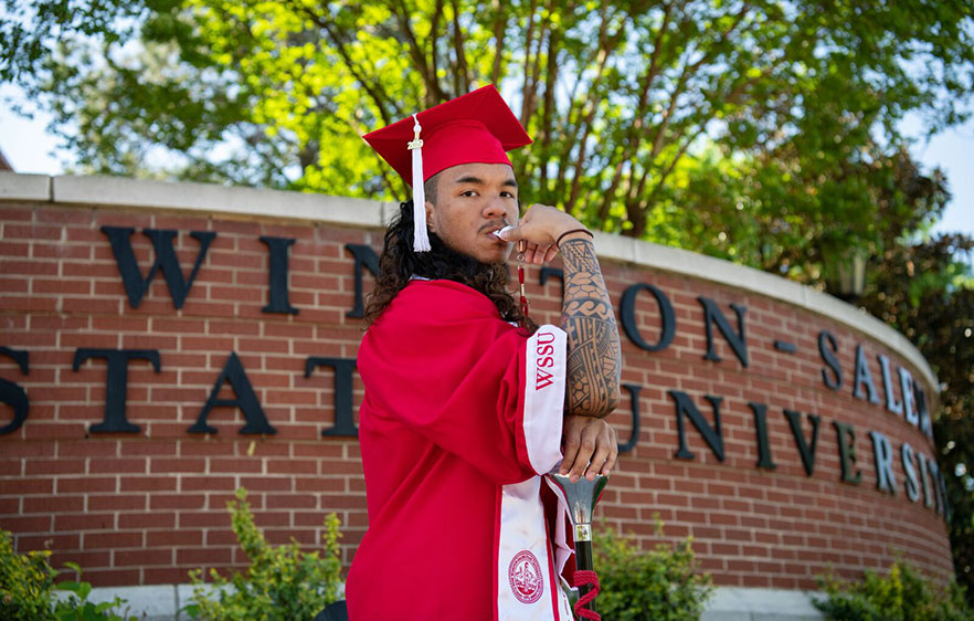 Greg Hughes flexes as he stands in cap and gown in front of the Winston-Salem State University sign on campus