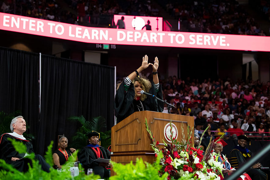 Kwanza Jones stands at the podium at WSSU's commencement, forming a W with her hands