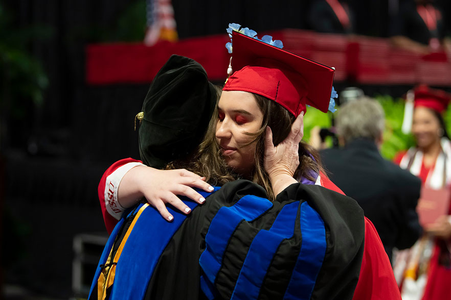 Student in red cap and gown hugs her mother, donning blue regalia